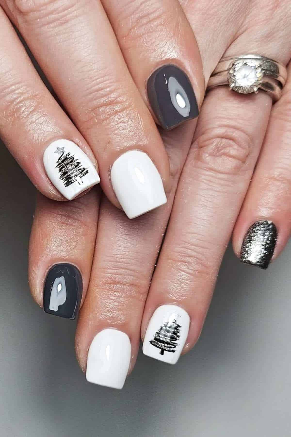 A hand with manicured nails featuring white, gray, and metallic silver polish, with black tree designs on two white nails.