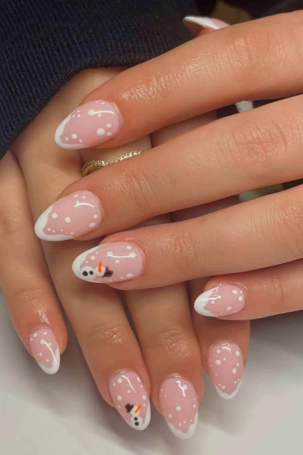 Hands with almond-shaped nails featuring a winter-themed manicure, including white tips, snow-like dots, and a snowman design on two accent nails.