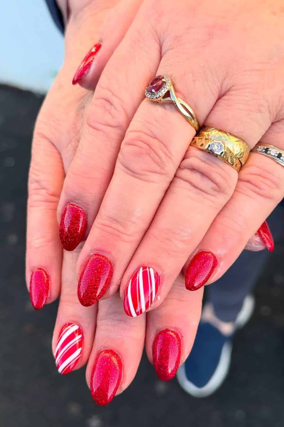 Hands with red glittery almond-shaped nails; two nails have red and white diagonal striped designs. The person wears multiple gold rings.