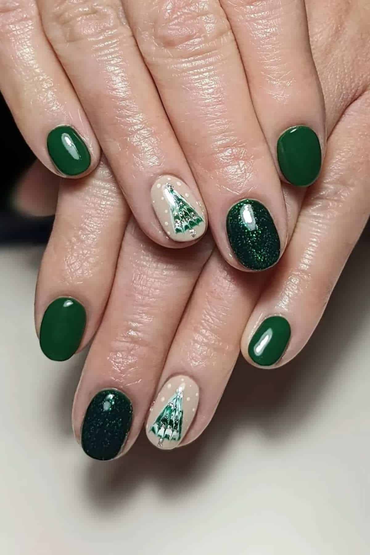 Hands with manicured nails painted in green and dark green polish, with accent nails featuring small Christmas tree designs on a light background.