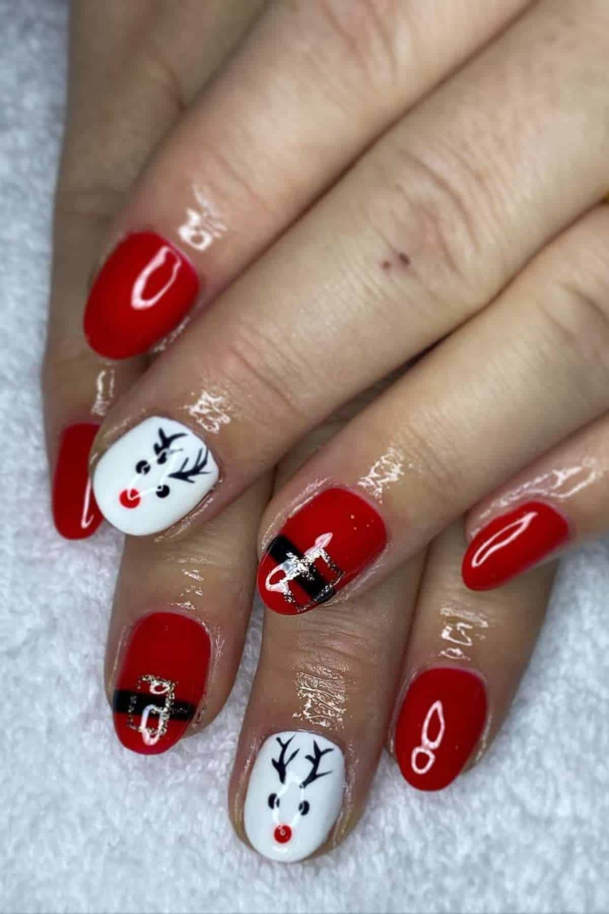 A hand with festive nail art featuring red polish, white accent nails with reindeer faces, and black belt designs resembling Santa's outfit.