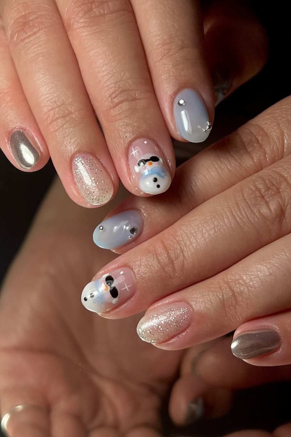 Hands with manicured nails featuring silver chrome, glitter, and snowman nail art designs on a light blue and nude base.