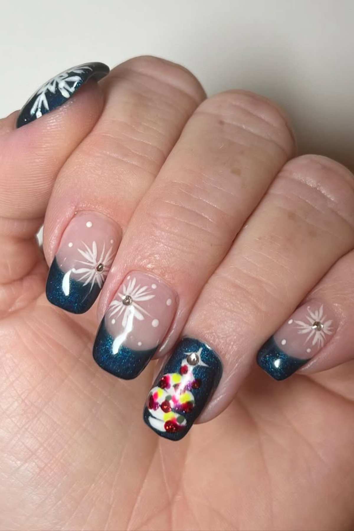 A hand with square-shaped nails featuring blue French tips, white snowflake designs, and a small, colorful decorated Christmas tree on one nail.