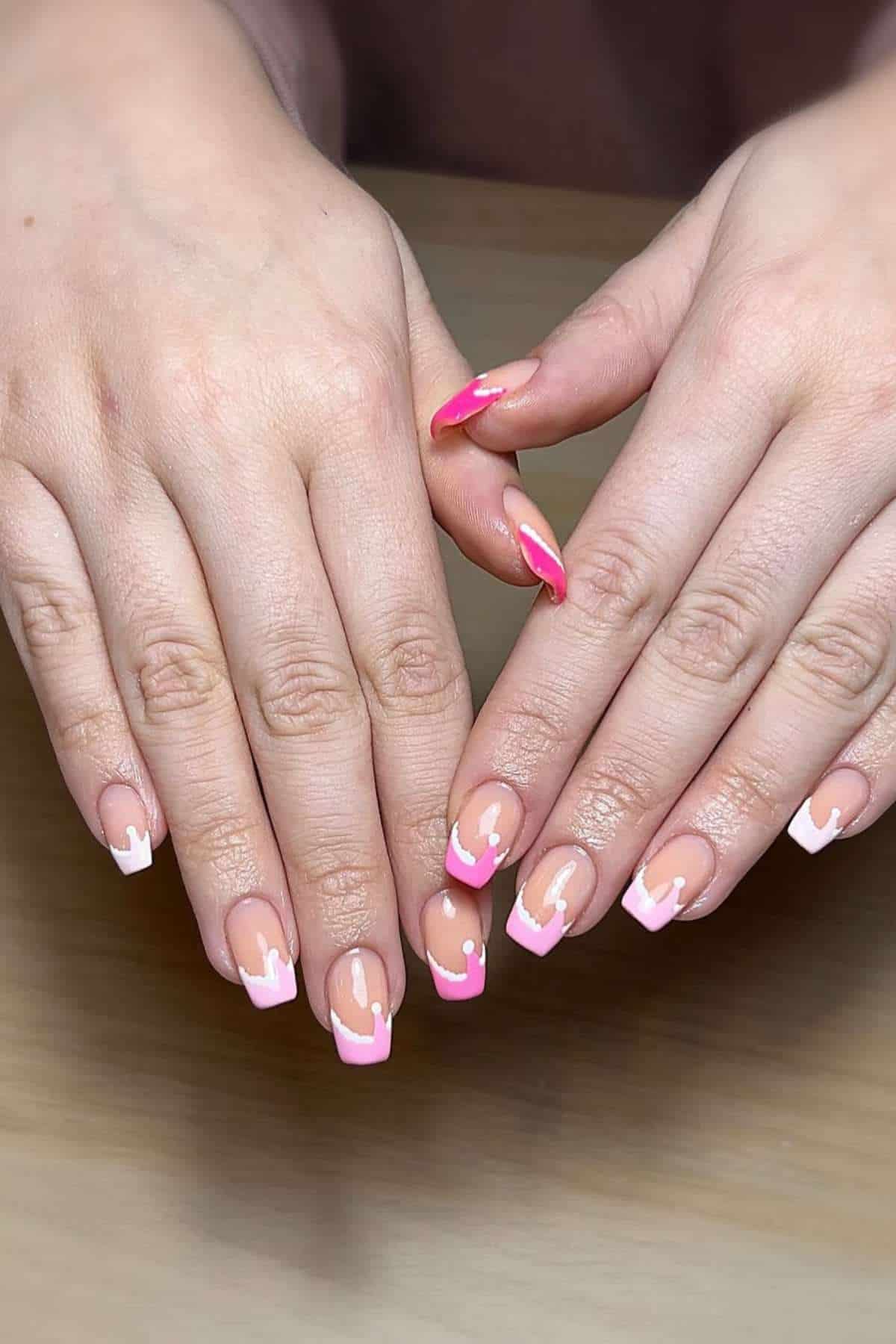 Hands with neatly manicured nails featuring a pink and white geometric French tip design, resting on a wooden surface.