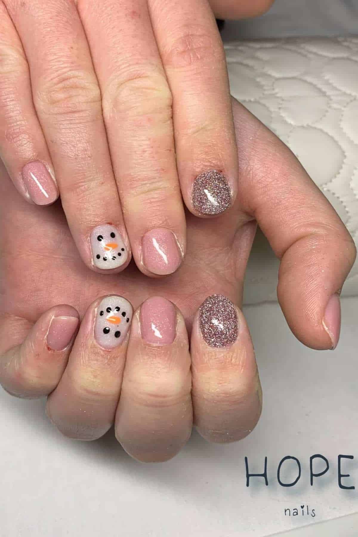 A hand with short, manicured nails features nude polish, glitter accent nails, and snowman faces painted on two nails. The background shows a quilted, light-colored surface.