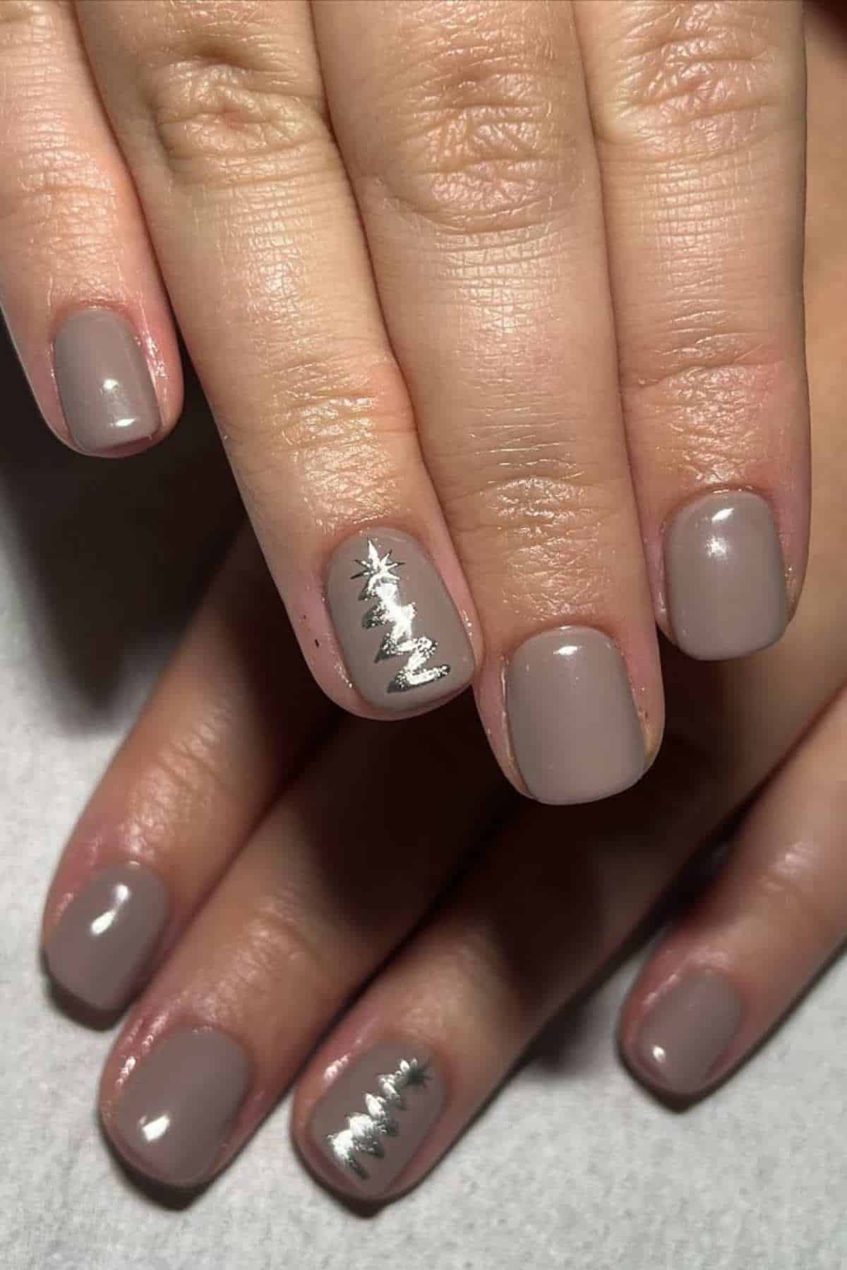 Hands with short, taupe-colored nails; one accent nail on each hand features a silver Christmas tree design.