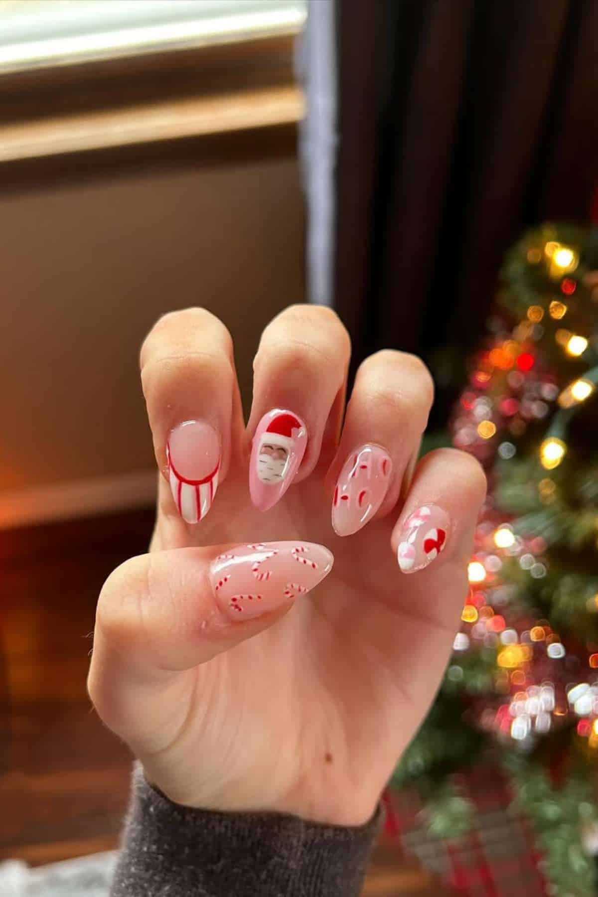 A hand with Christmas-themed nail art including candy canes, Santa’s face, red and white patterns, held up near a decorated Christmas tree.