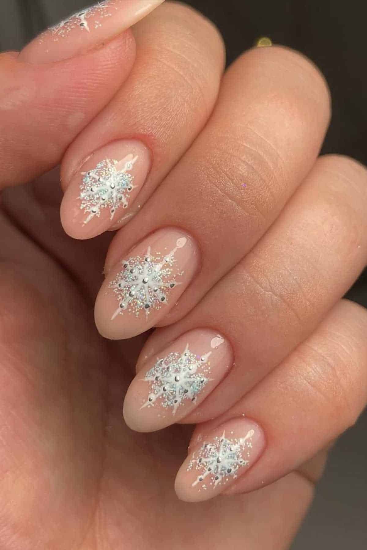 A close-up of a hand with almond-shaped nails painted nude and decorated with white and silver snowflake designs.