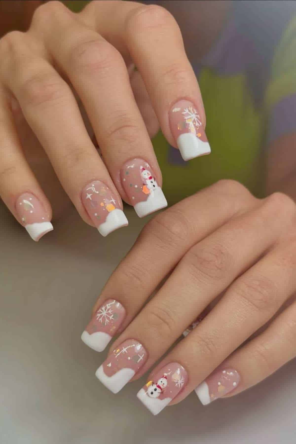 Hands with French manicured nails featuring holiday-themed designs, including snowflakes, stars, and snowmen on a light pink base.