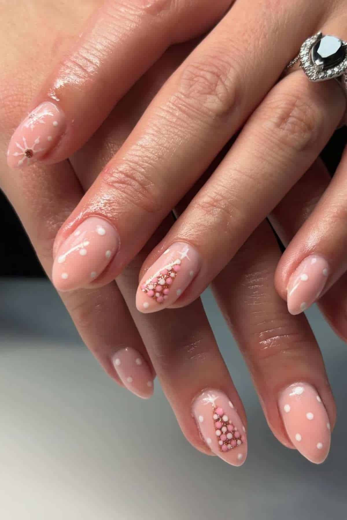 A hand with almond-shaped nails painted light pink, featuring white polka dots and small pink and white bead designs, next to a silver ring with a black stone.