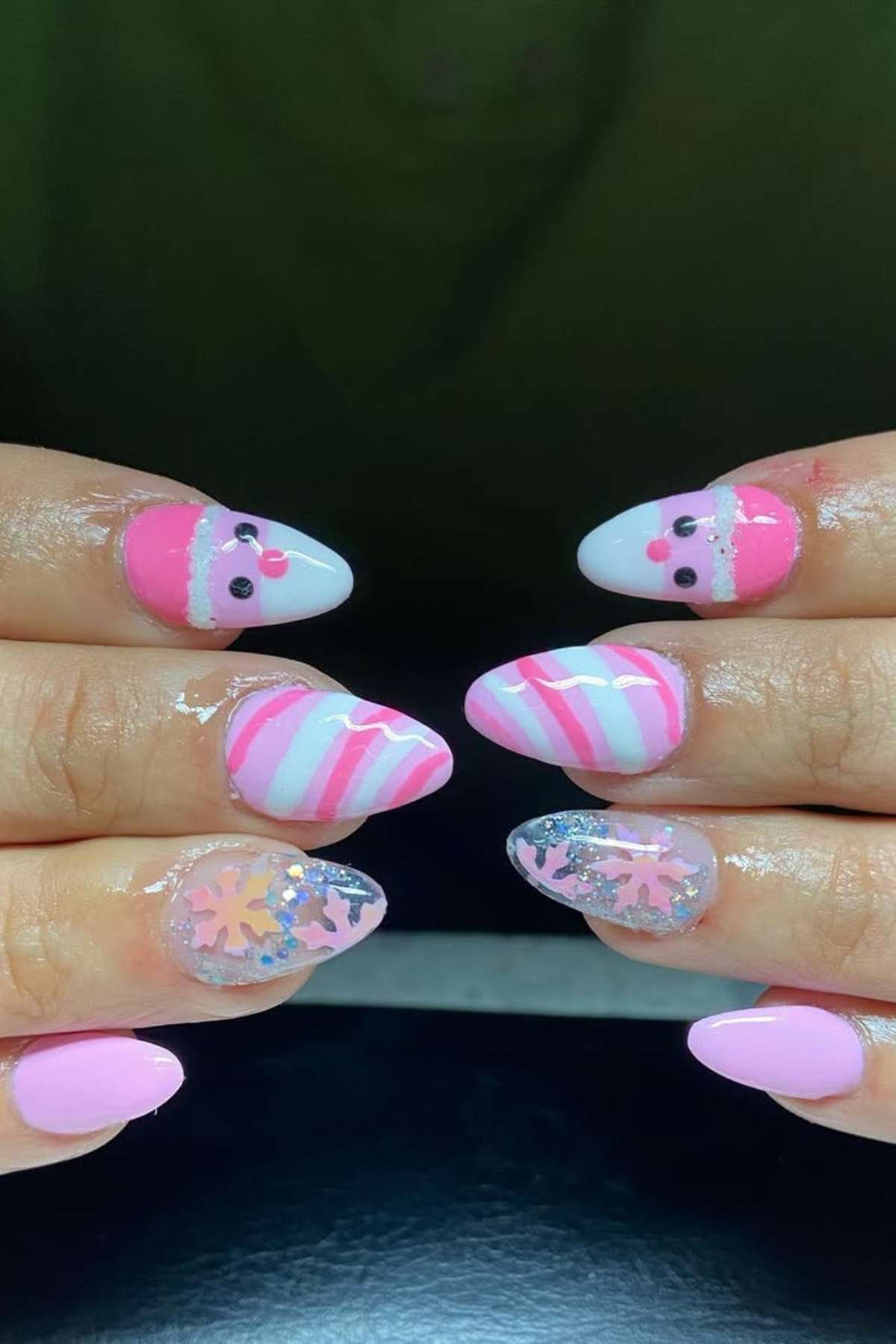 Hands with almond-shaped nails featuring pink and white Santa faces, striped patterns, glitter snowflakes, and solid pink polish, displayed against a dark background.