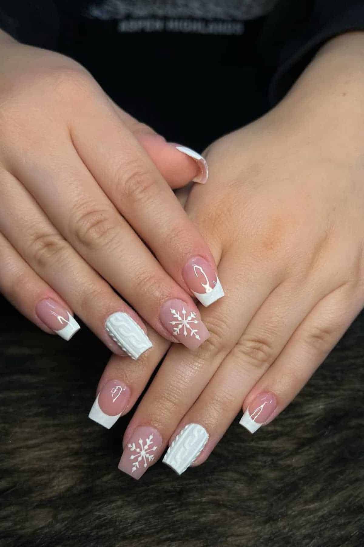 Hands with manicured nails featuring white French tips, white snowflake and cable knit designs on a few nails, displayed on a dark surface.