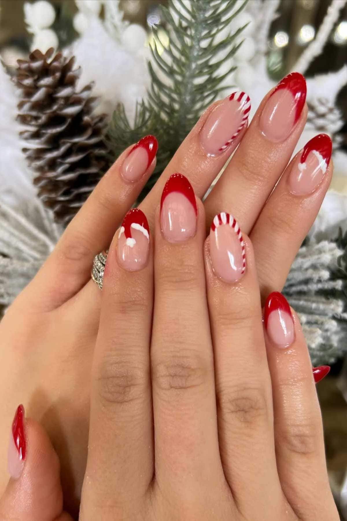 Hands with almond-shaped nails featuring a red French tip design, with some nails accented by red and white candy cane stripes, against a background of pine cones and greenery.