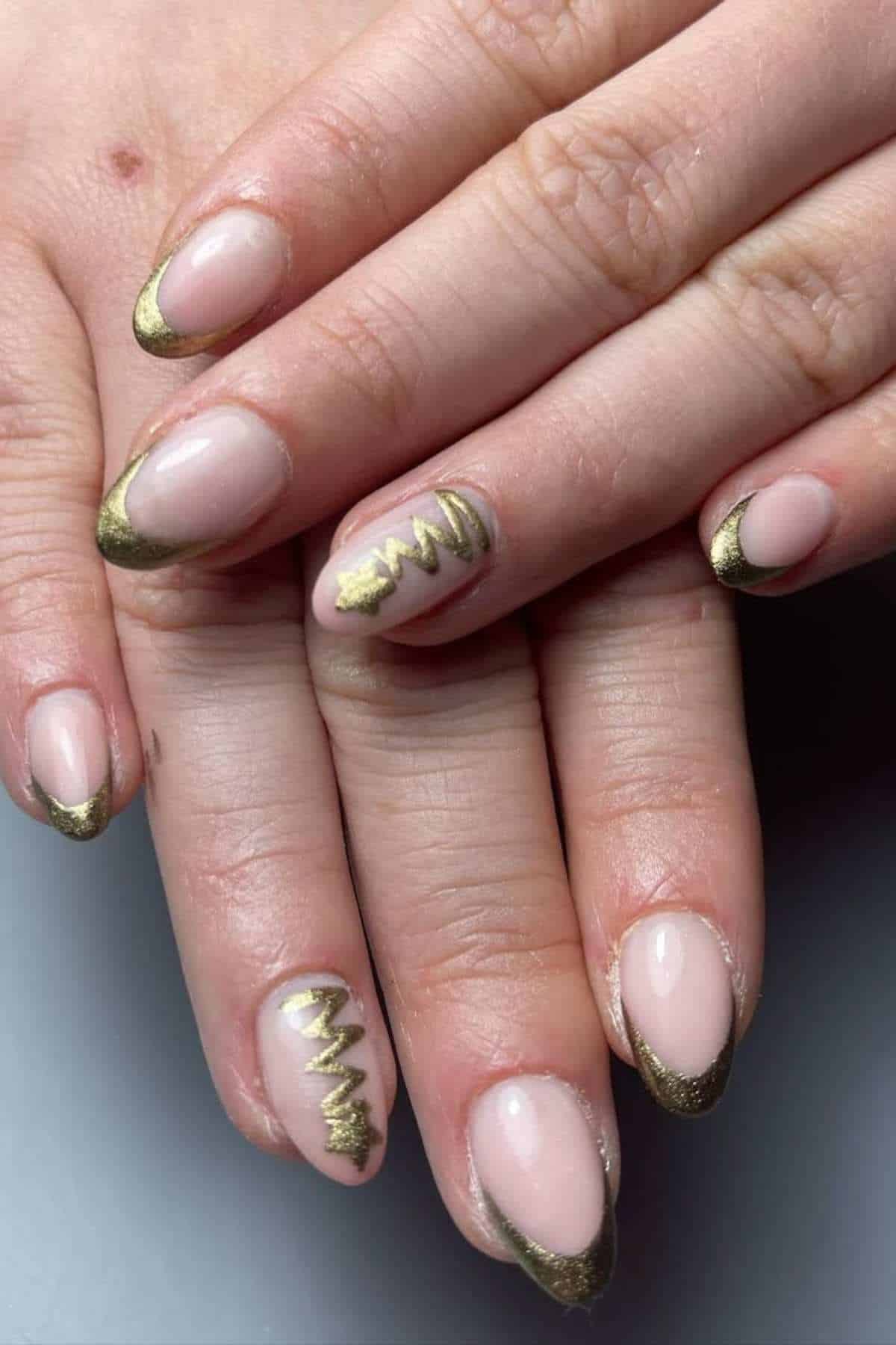 Hands with almond-shaped nails painted nude with metallic gold tips; two nails feature a gold Christmas tree design.