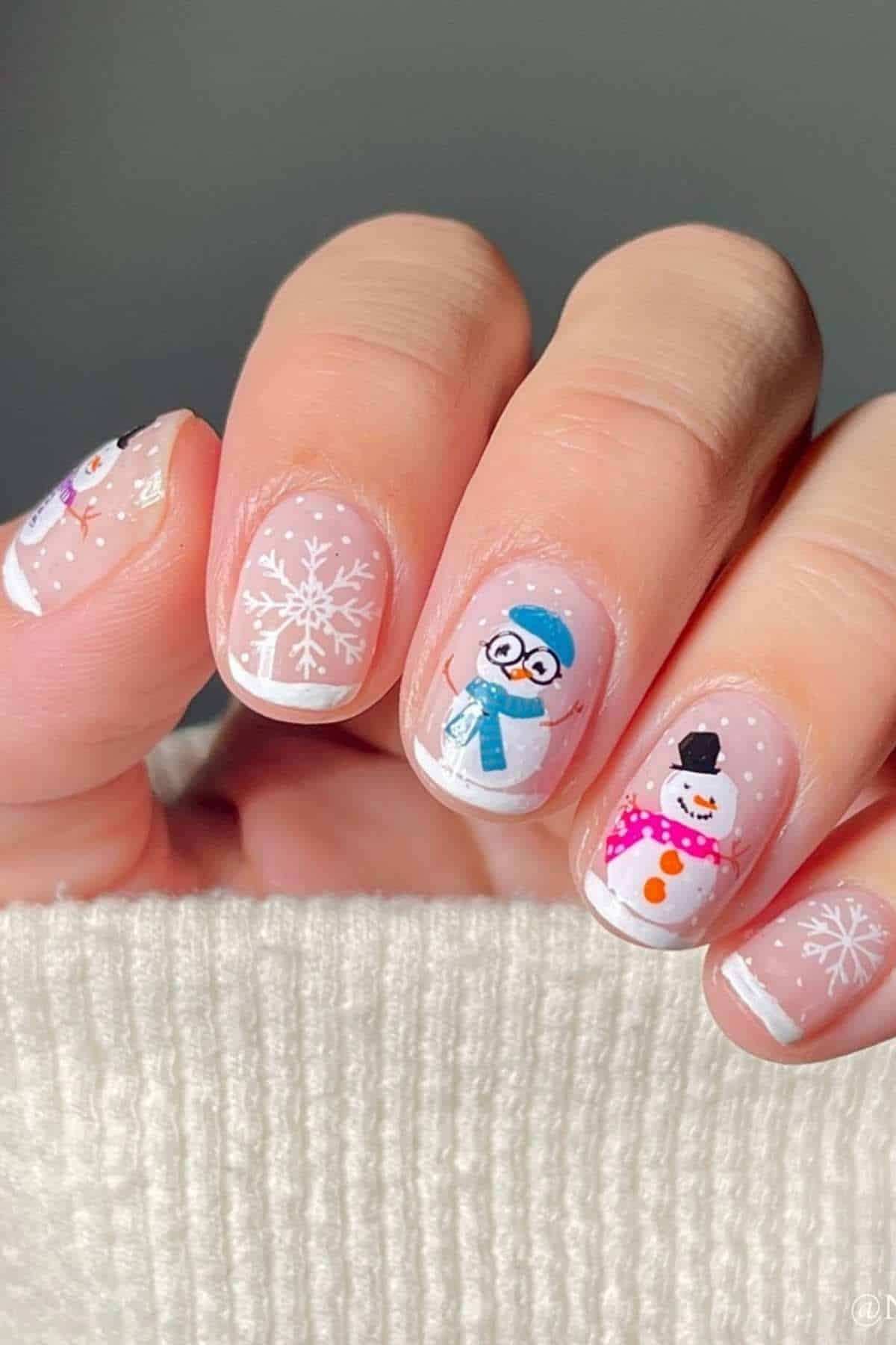 A hand with winter-themed nail art featuring white snowflakes and cartoon snowmen on a light pink base.