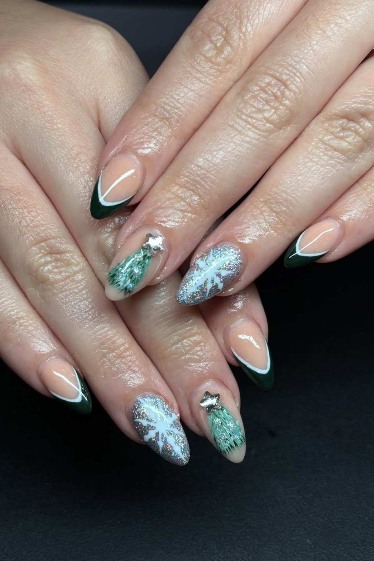 Hands with almond-shaped nails featuring green French tips, glitter accents, and intricate holiday-themed designs such as Christmas trees and snowflakes.