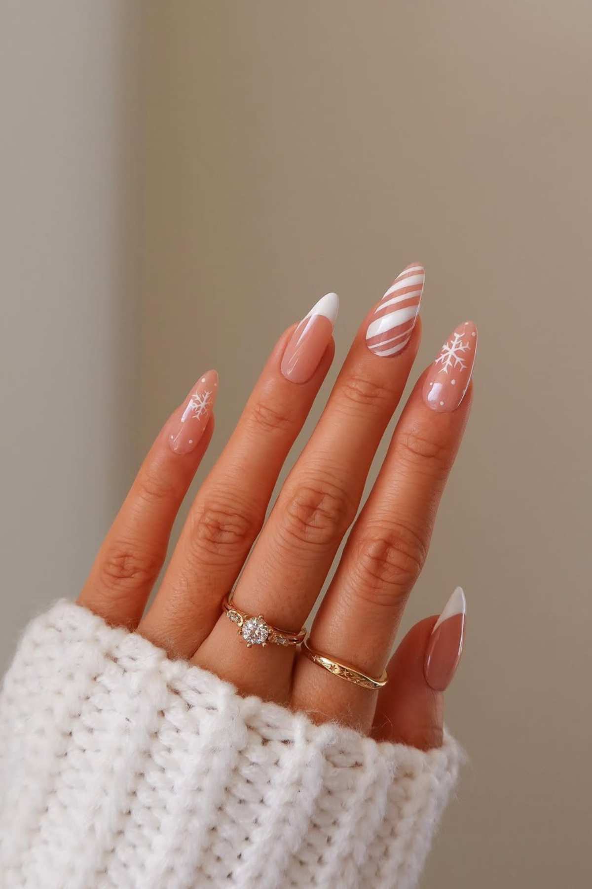 A hand with long, almond-shaped nails featuring nude polish, white tips, and festive white designs, wearing two gold rings and a white knit sweater.