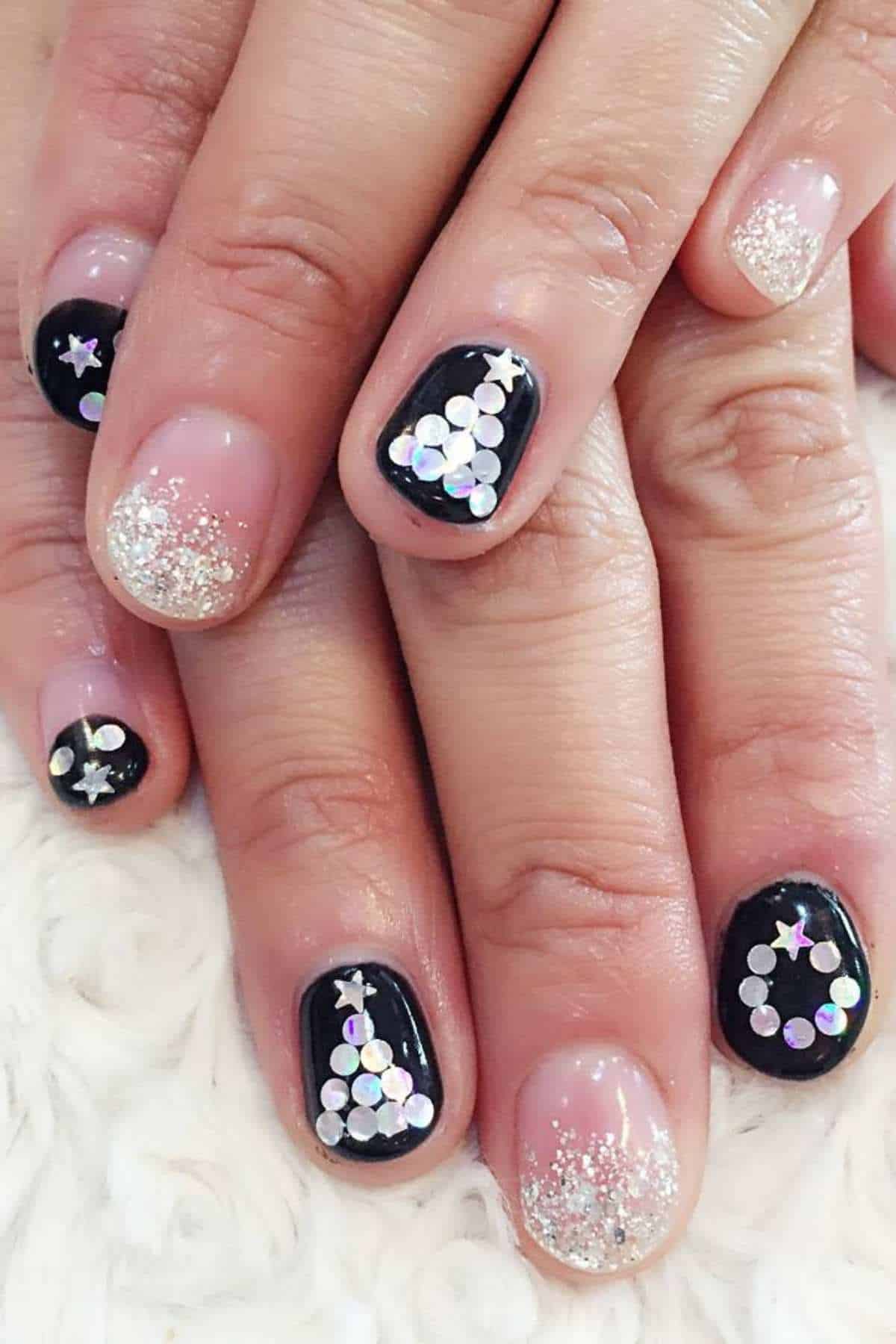 Hands with short nails featuring black and nude polish, silver glitter tips, and holographic sequin decorations forming circles and stars on some nails.