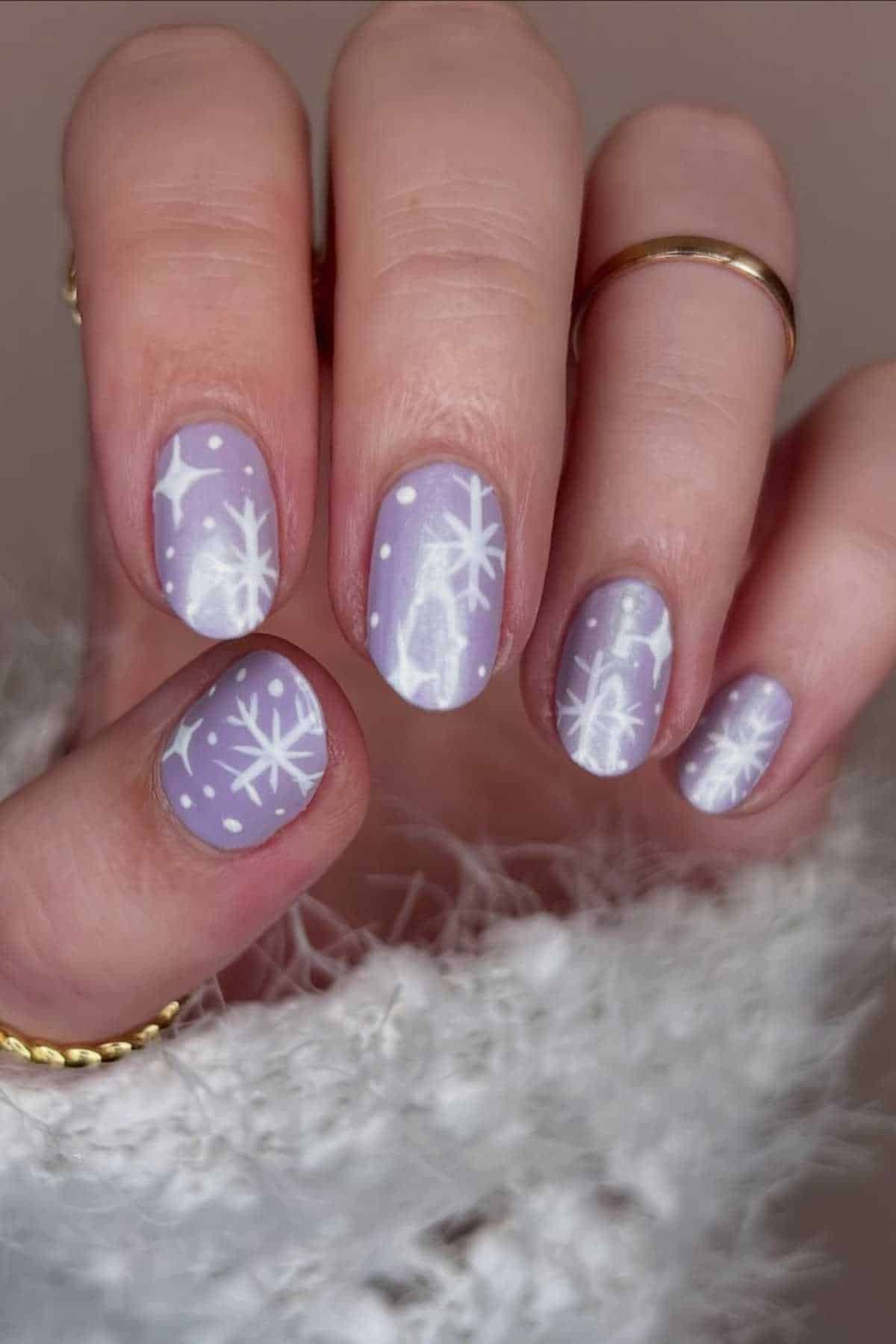 A hand with lavender nail polish featuring white snowflake and dot designs, wearing gold rings and a white fuzzy sweater.
