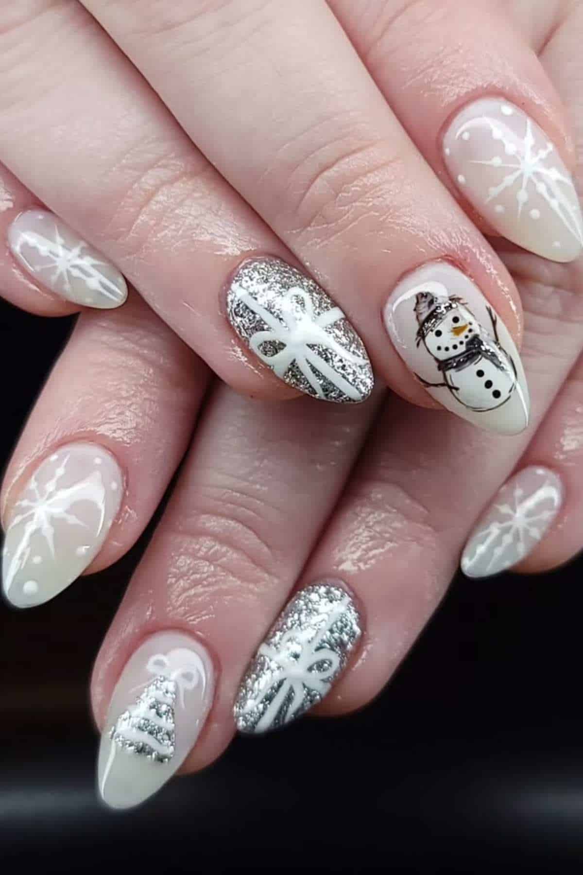 Close-up of hands with almond-shaped nails featuring white snowflakes, a snowman, Christmas trees, and silver glitter designs on a neutral base.