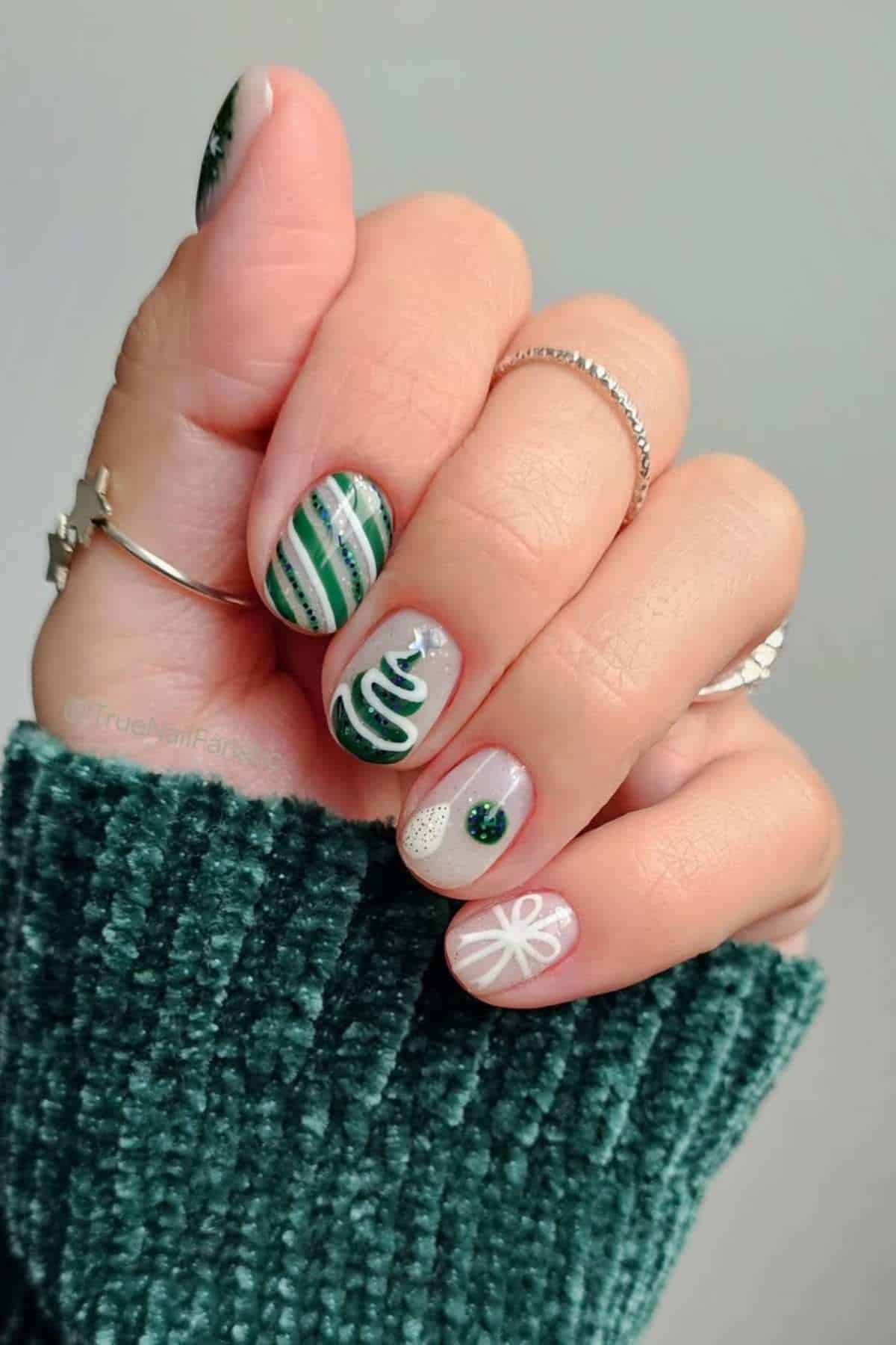 A hand with short nails painted in green and white Christmas-themed designs, including a tree, stripes, dots, and a bow, wearing silver rings and a green sweater.