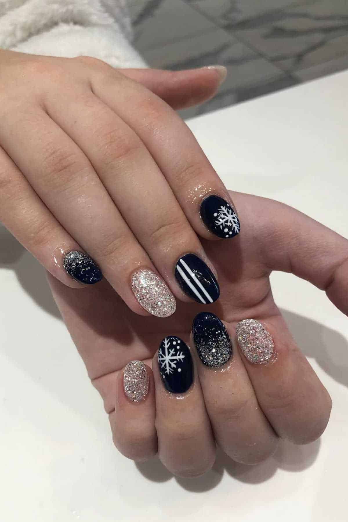 Hands showing nails painted with a mix of black and glitter polish, featuring snowflake designs and a white stripe accent.