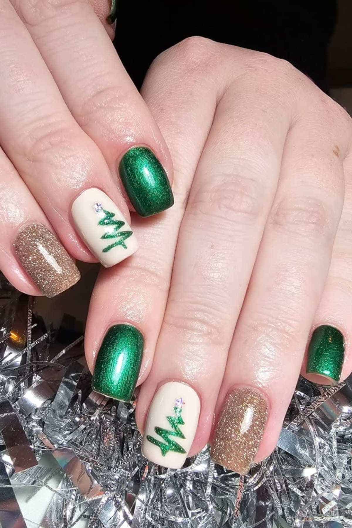 Hands with manicured nails featuring green, gold glitter, and cream polish; two nails have Christmas tree designs with small decorations.