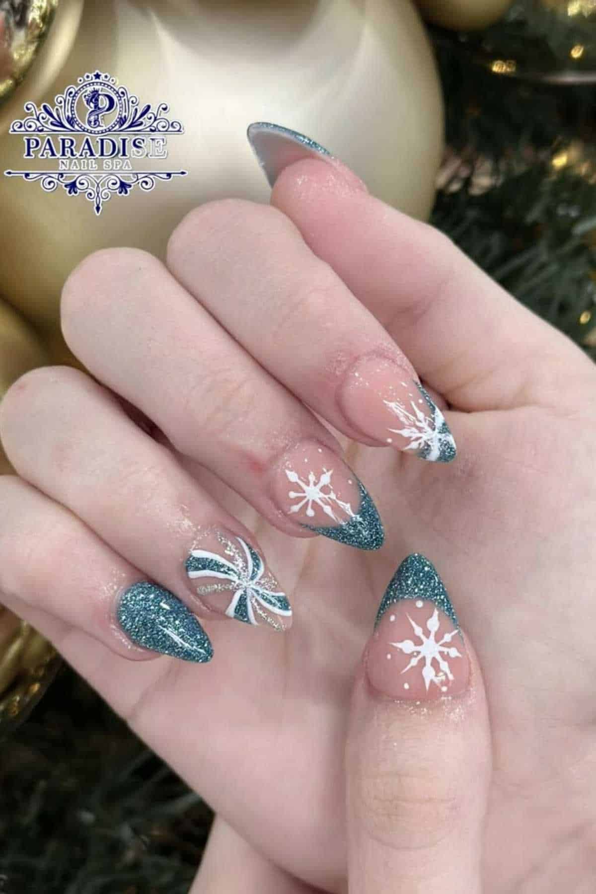 A hand with teal glittery stiletto nails featuring snowflake and pinwheels designs, photographed near a Christmas tree.