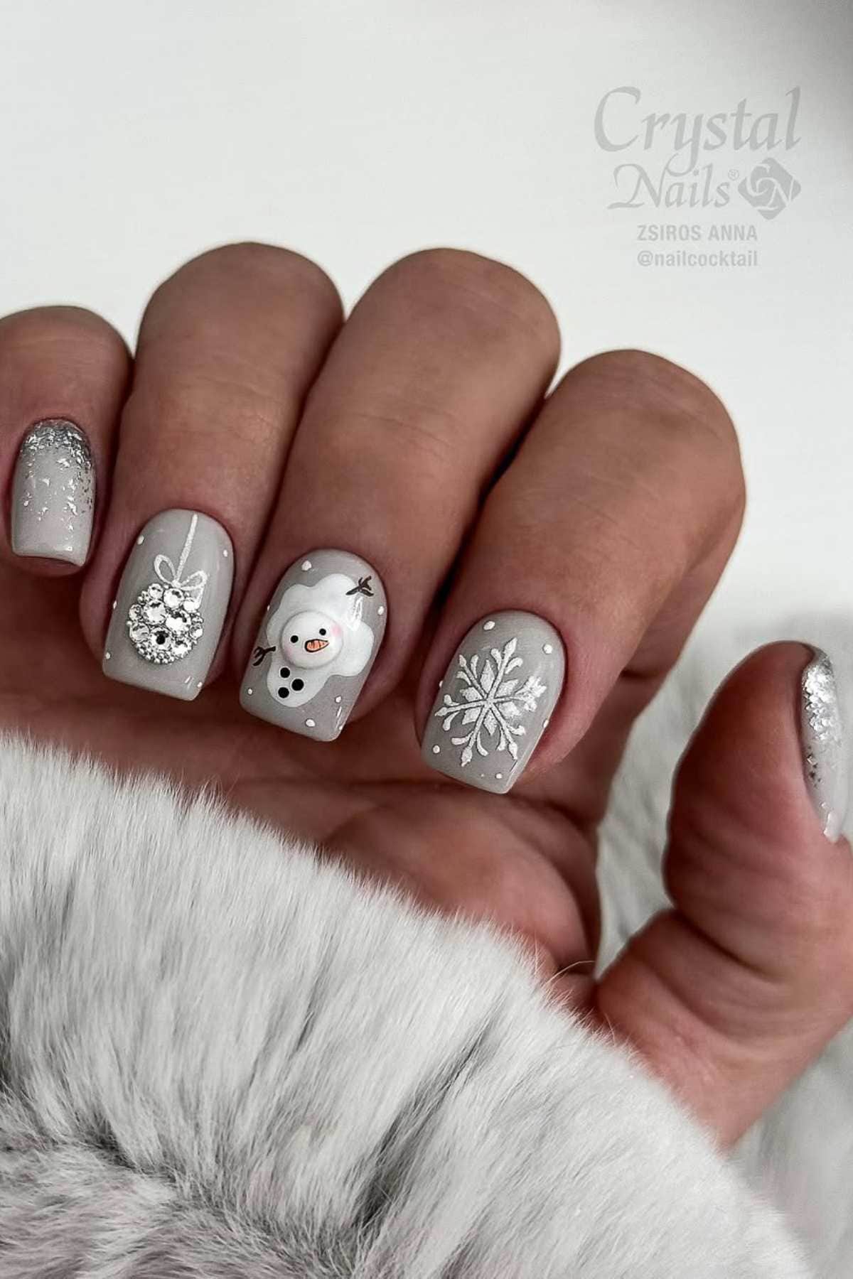 A hand with short, square-shaped nails painted light grey, featuring designs of a snowflake, a snowman, glitter, and white accents, resting on a fur-like fabric.
