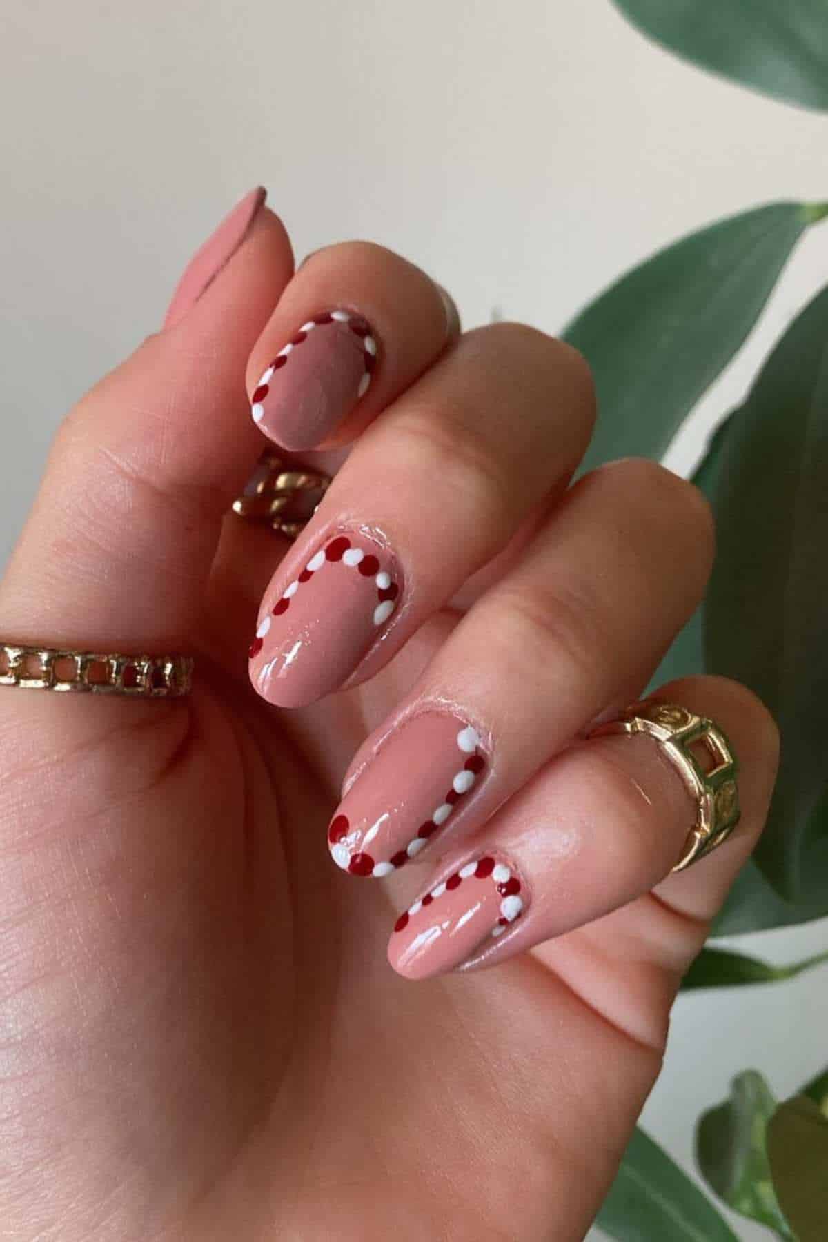 A hand with nude pink nail polish features a dotted red and white design along the cuticles; gold rings are worn on the fingers.