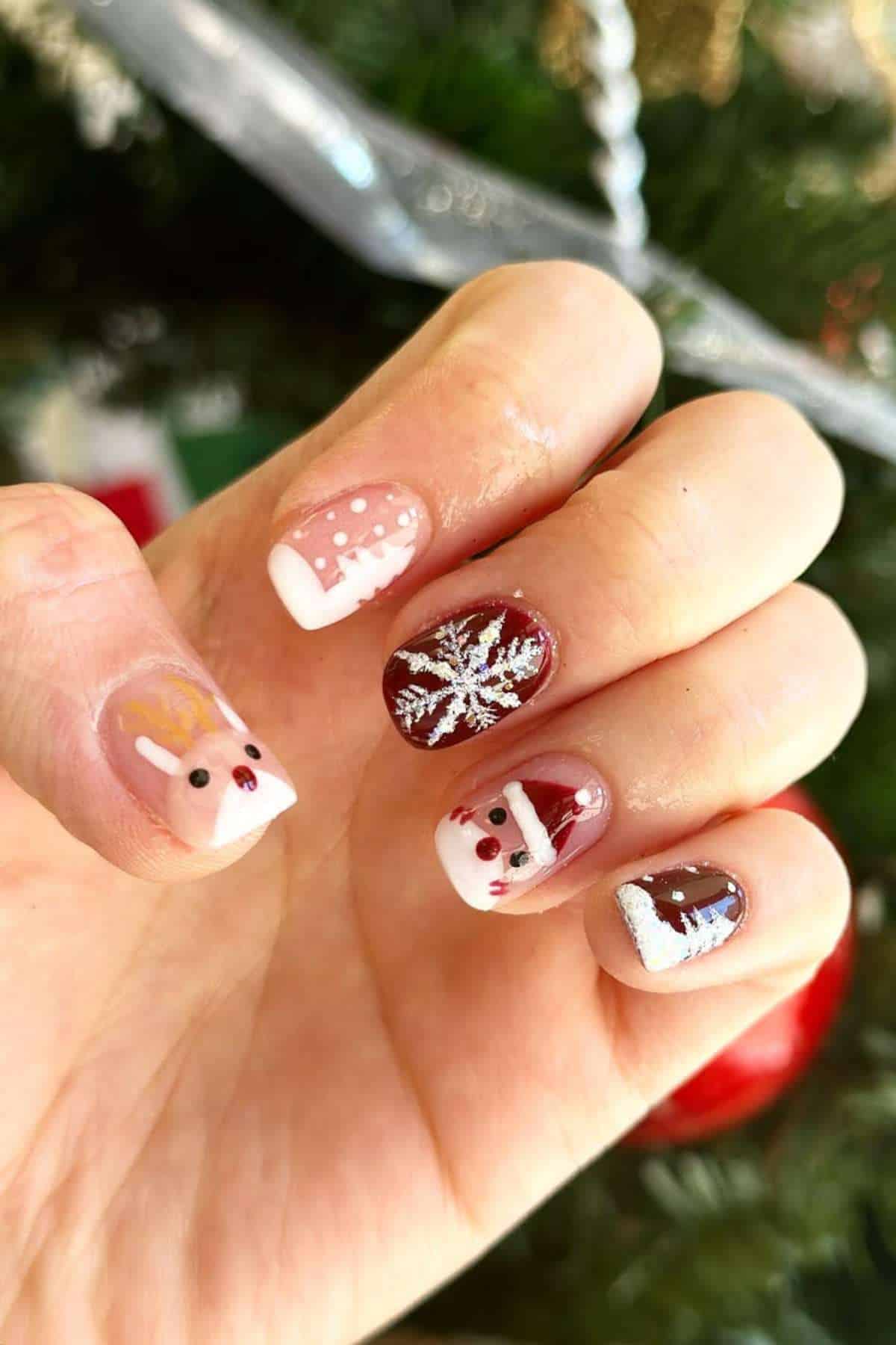 A hand with festive Christmas nail art, including designs of a reindeer, snowflakes, Santa Claus, and a Santa hat against a blurred holiday background.