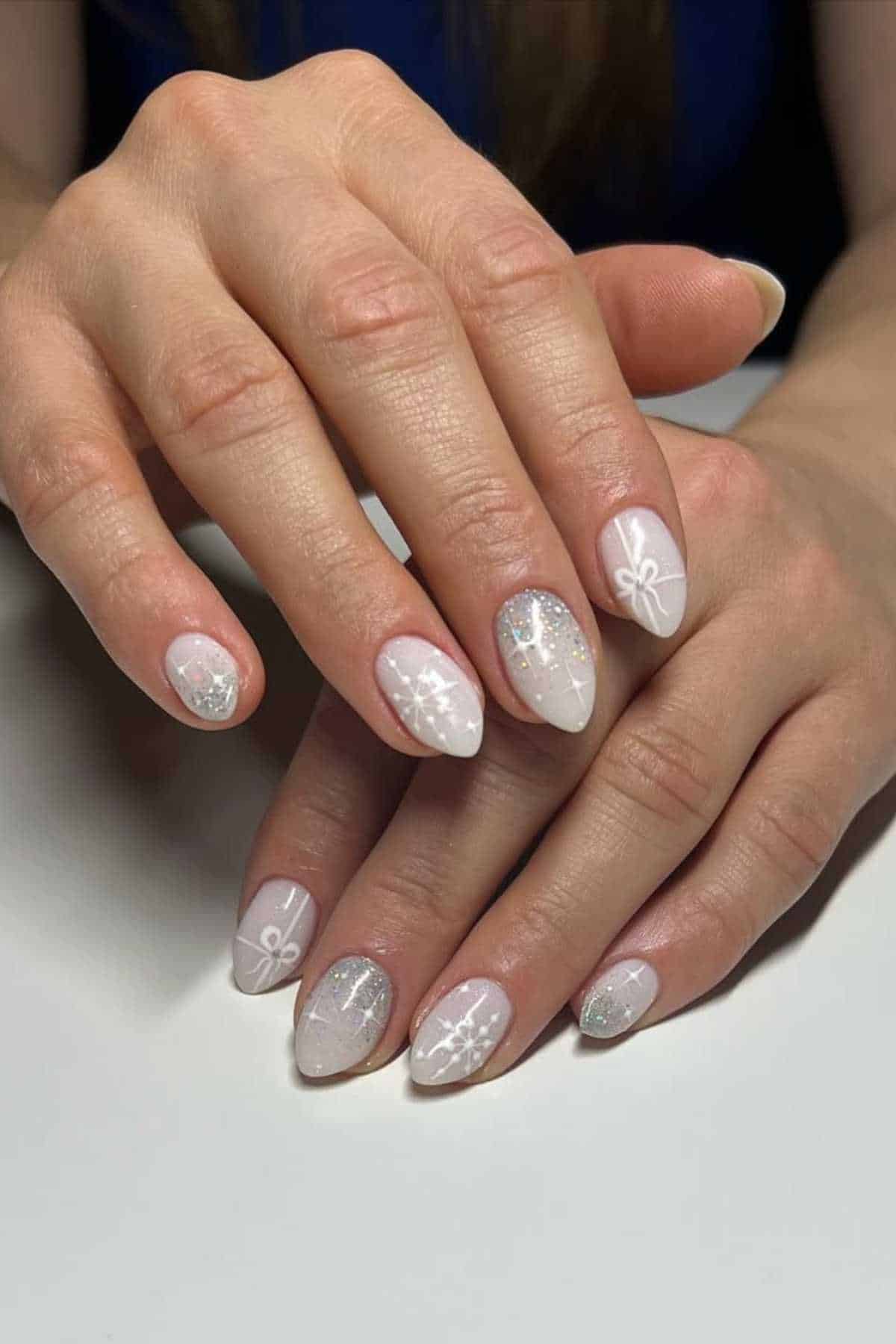Hands with almond-shaped nails painted light pink, featuring white bow and snowflake designs, and some nails with silver glitter accents.