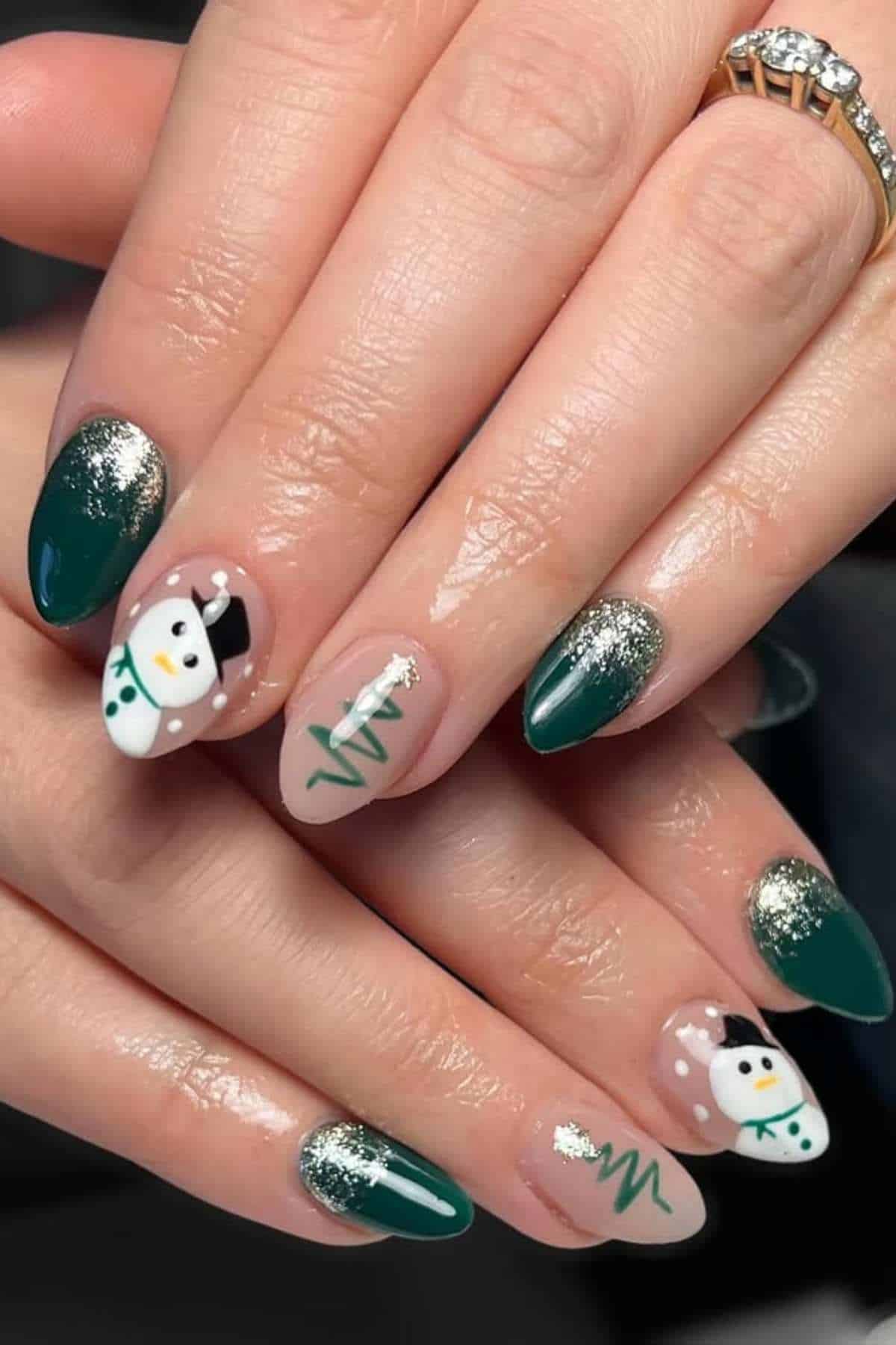 Hands with green, gold, and nude nail polish featuring snowman and Christmas tree designs; person is wearing a gold ring with a clear stone.