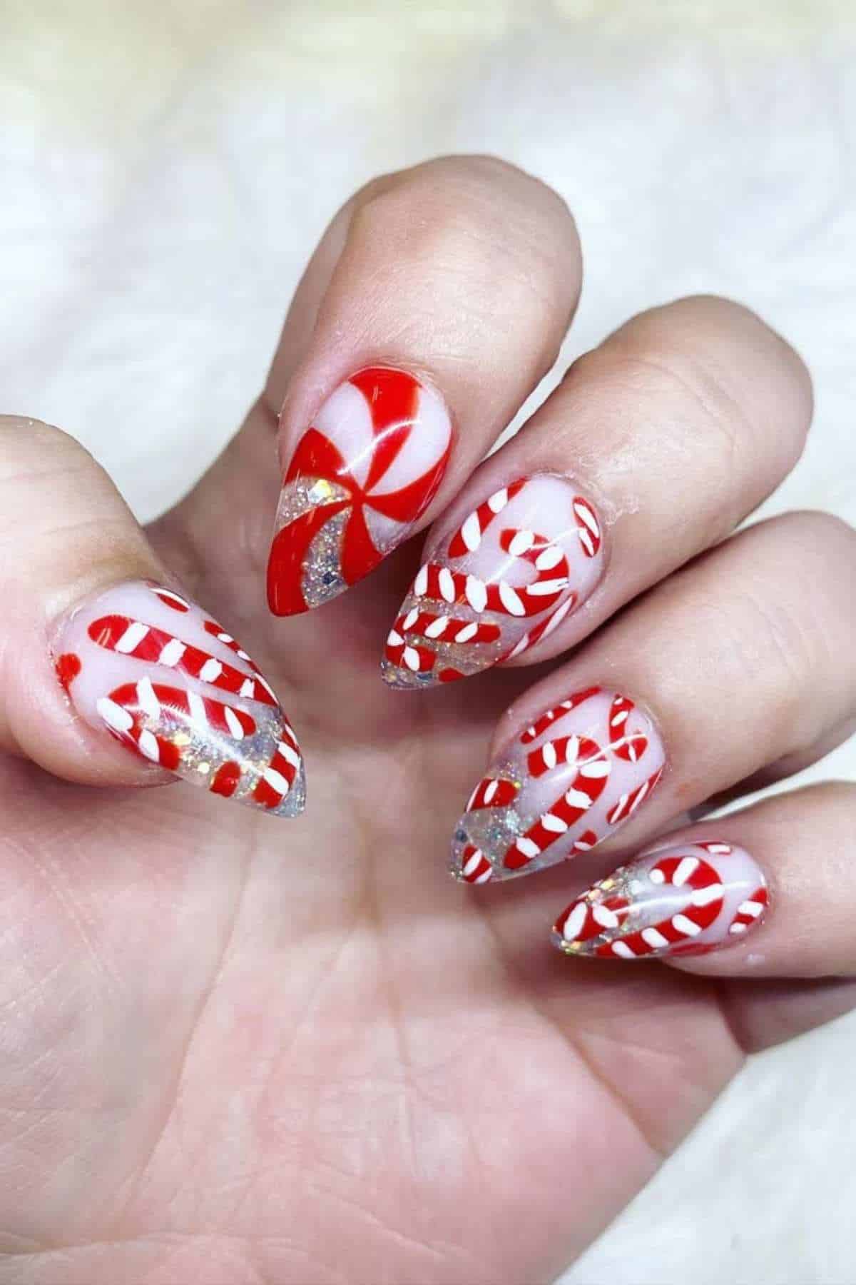 A hand with long, almond-shaped nails featuring red, white, and silver peppermint candy and swirl designs on each nail.