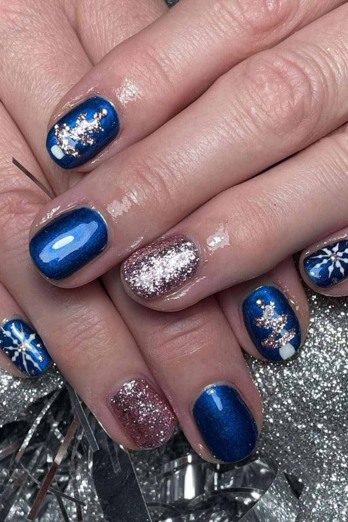 Hands with blue and glittery pink nail polish, featuring snowflake and Christmas tree designs, resting on silver tinsel.