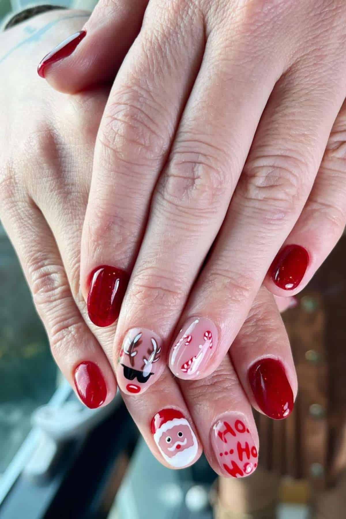 Hands with red and nude Christmas-themed nail art, including designs of a reindeer, candy canes, Santa face, and "Ho Ho Ho" text.