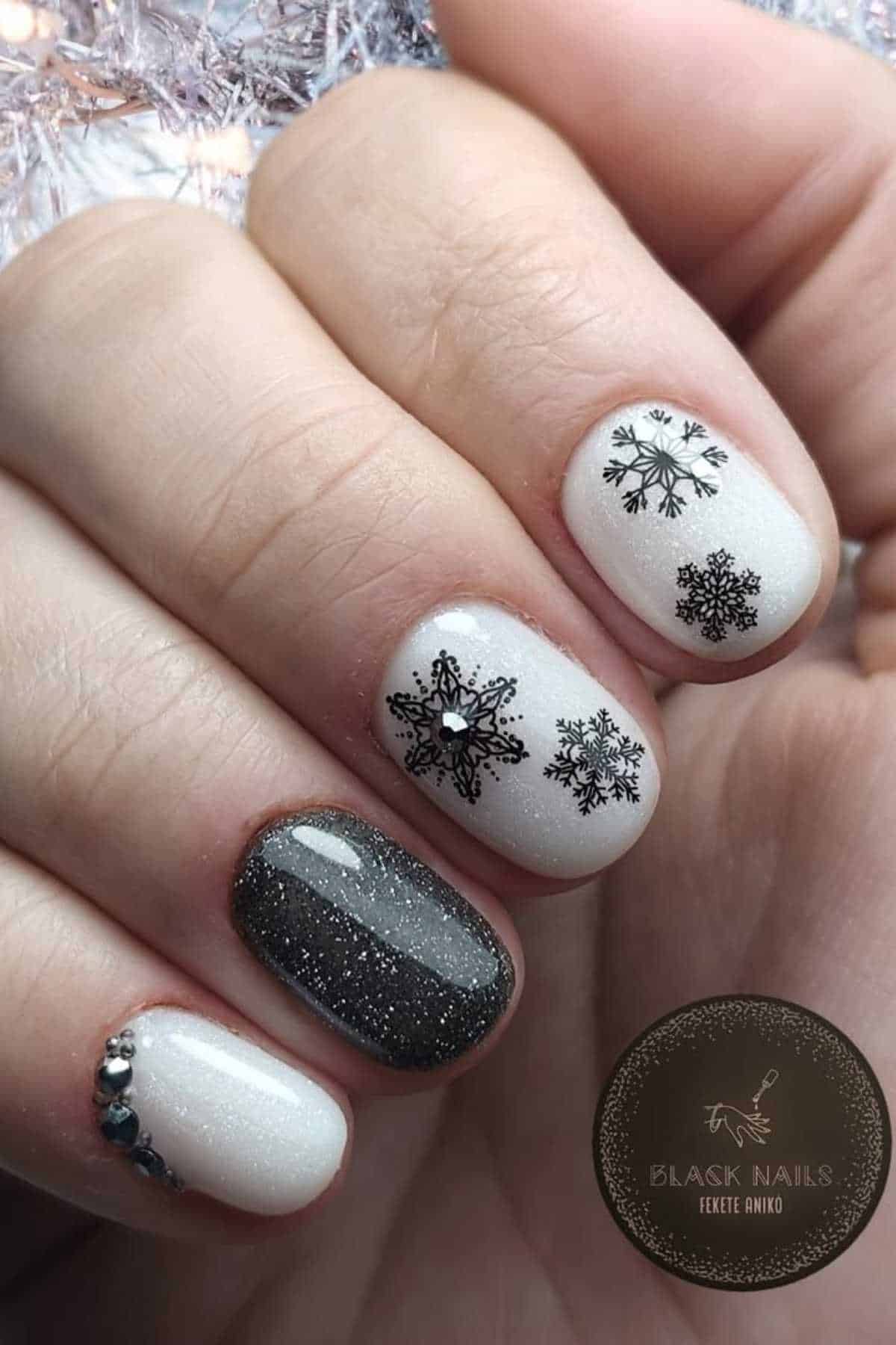 A hand with white and black winter-themed nail art featuring snowflakes, glitter, and small rhinestone accents.