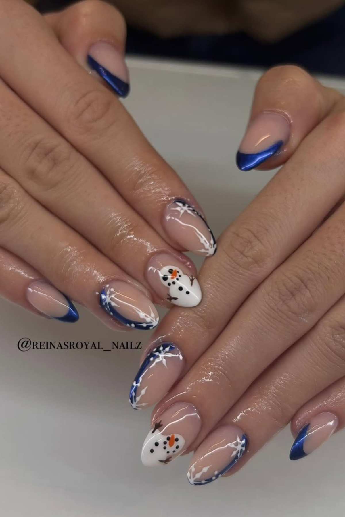 Hands with almond-shaped nails featuring blue French tips, snowflakes designs, and two accent nails with white bases and snowmen.