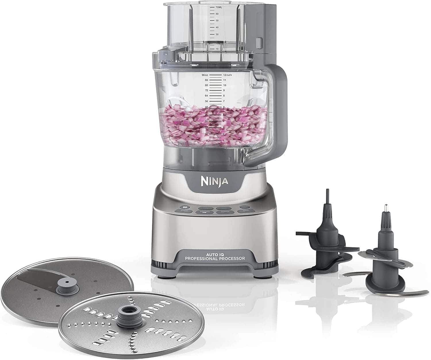 A Ninja professional food processor with a transparent container partially filled with chopped red onions, two blade attachments, and two slicing discs placed nearby.