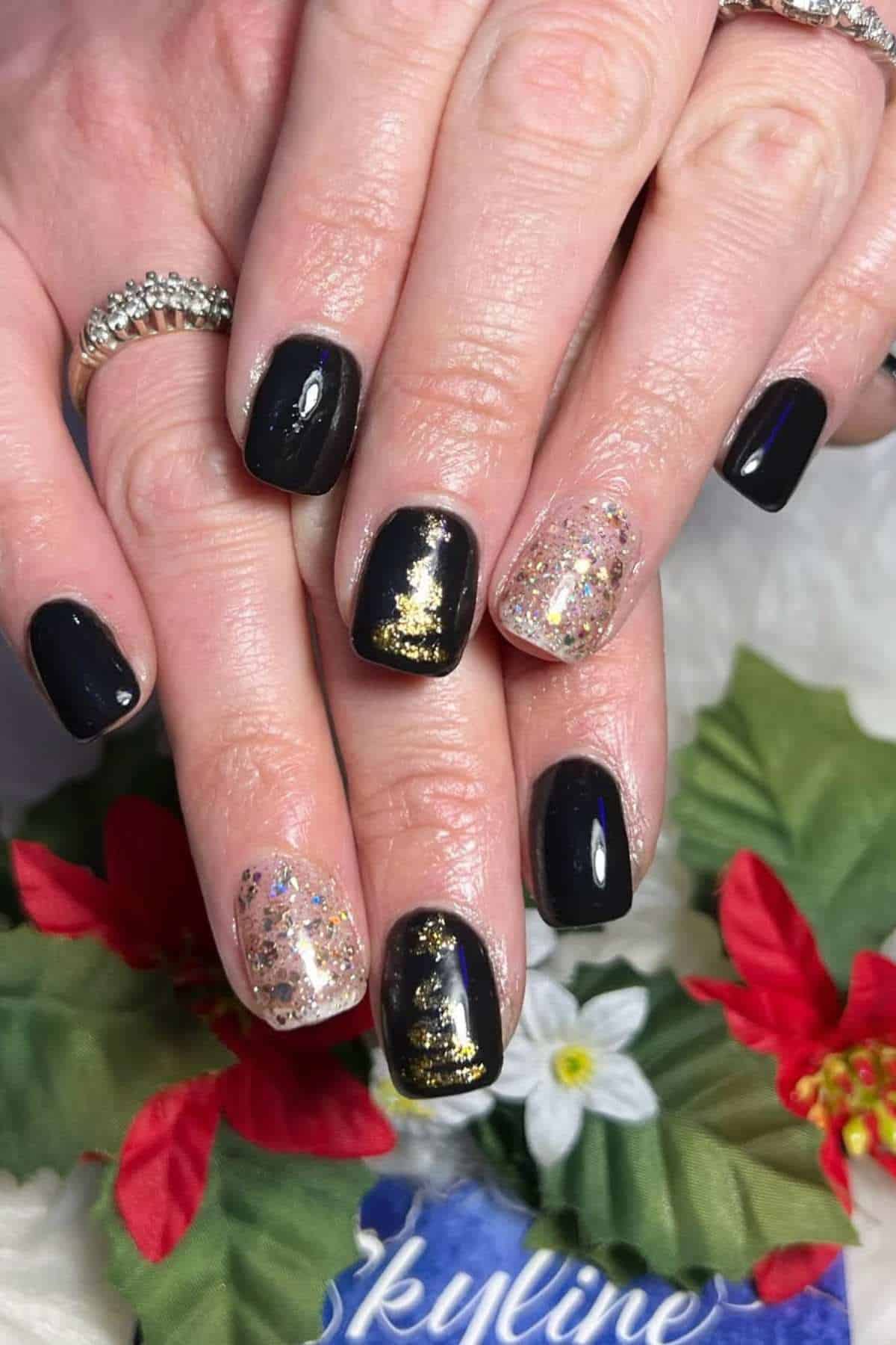 Close-up of hands with short nails painted black, with accent nails featuring gold foil and glitter. The hands are resting on a floral background.