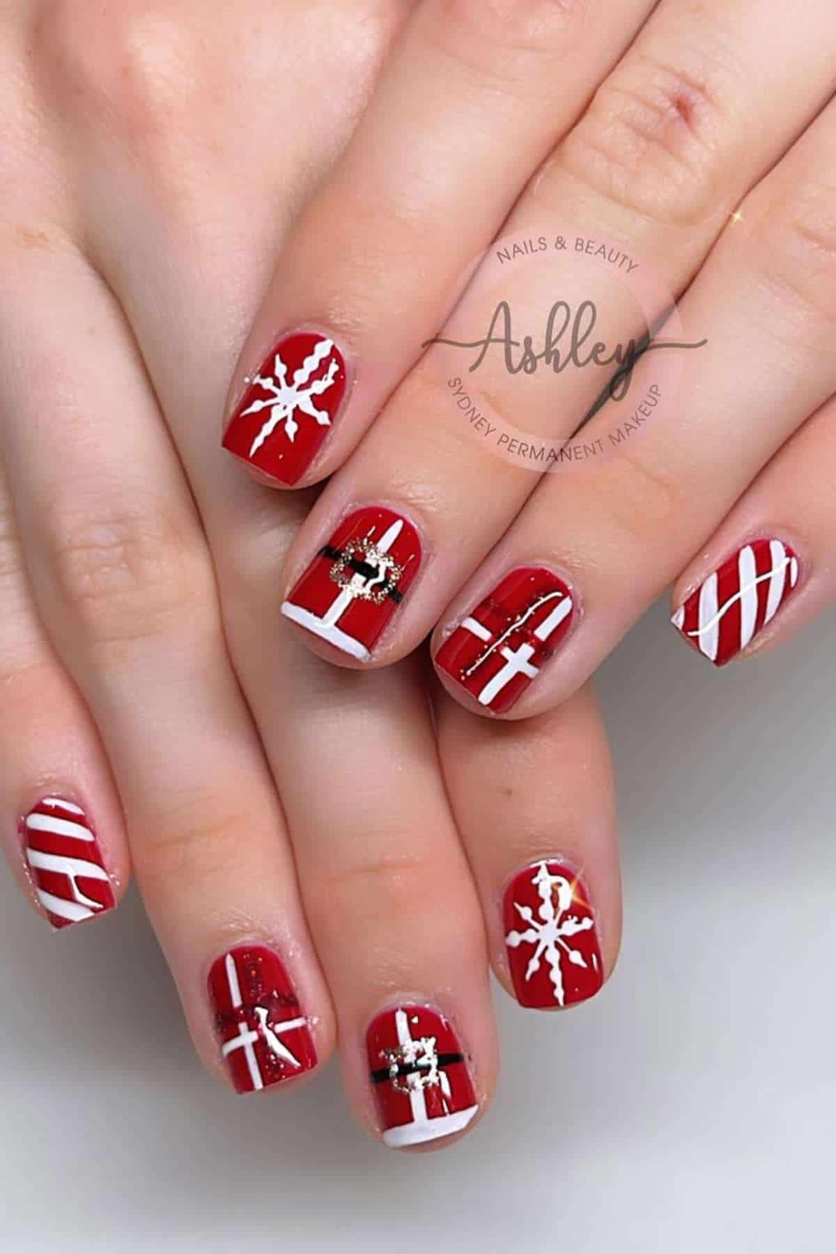 Hands with red manicured nails featuring white snowflakes, stripes, and gift bow designs, along with a gold embellishment accent.