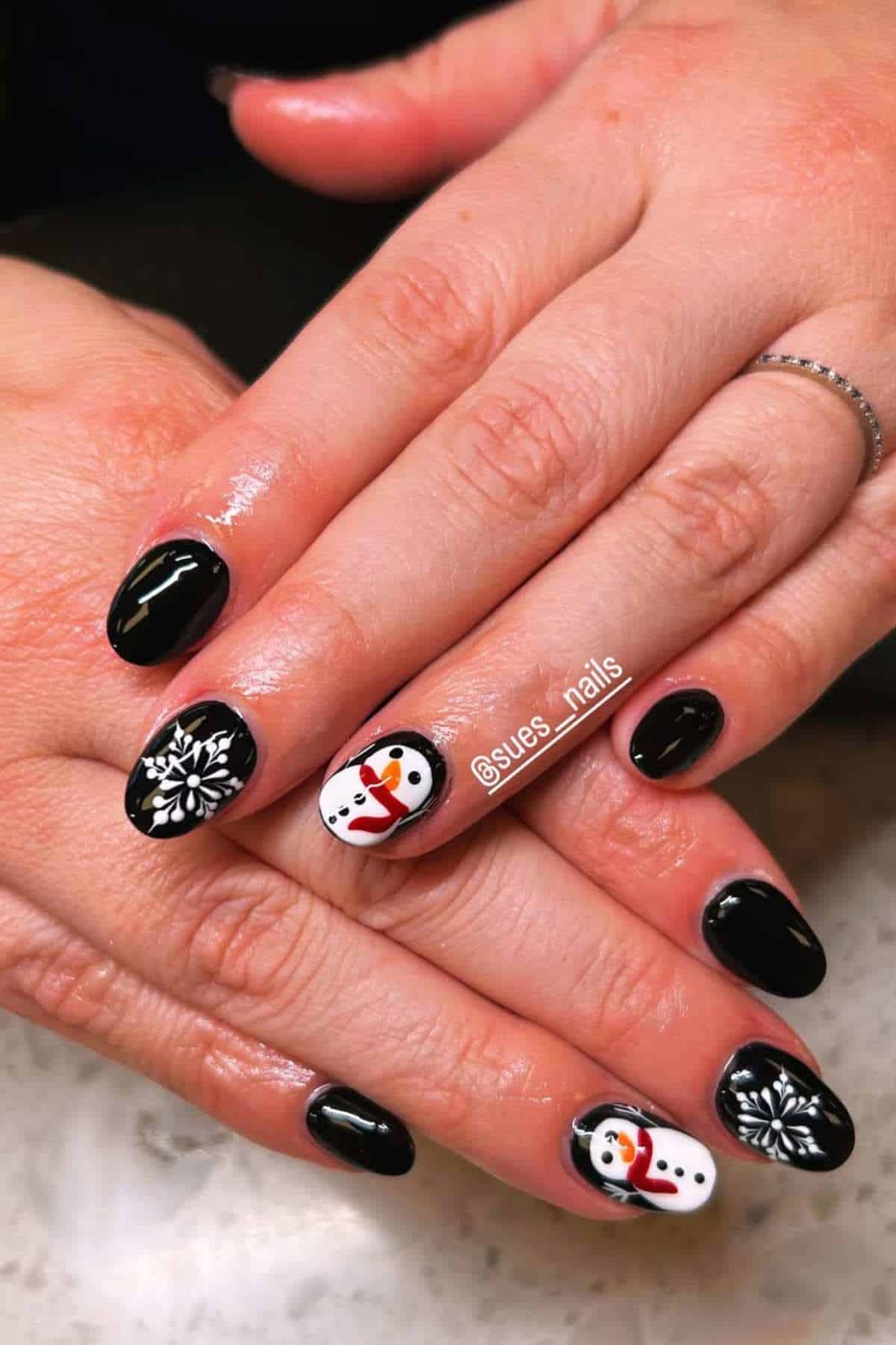 Hands with black nail polish, featuring accent nails with a snowflake design and a snowman illustration on a white background.