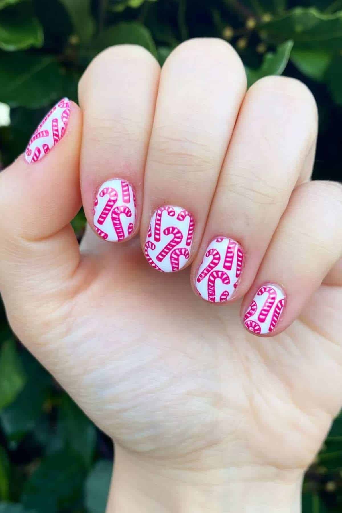 Close-up of a hand with fingernails painted white and decorated with pink candy cane patterns, held in front of green leafy background.