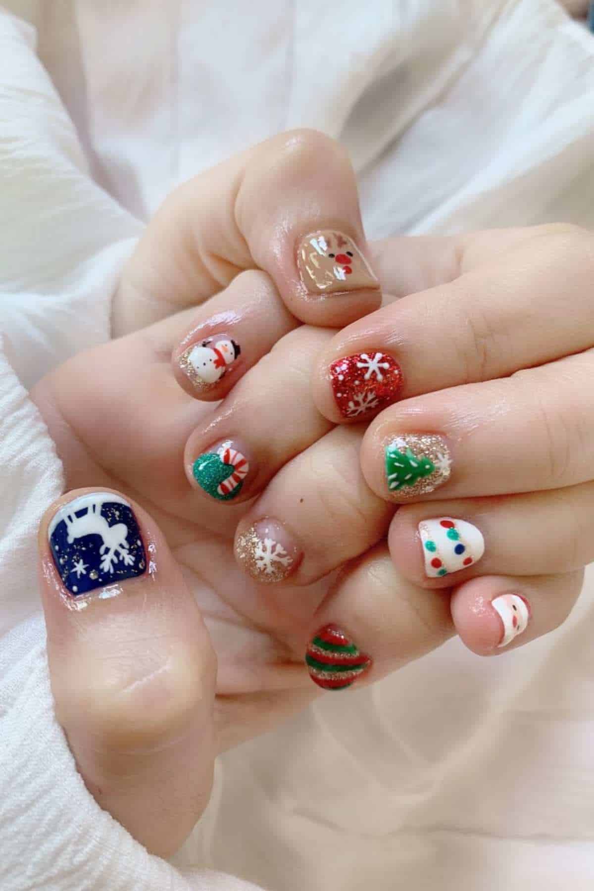 Hands with short nails painted in various Christmas-themed designs, including a reindeer, snowflakes, snowman, Christmas tree, ornaments, and holiday lights.