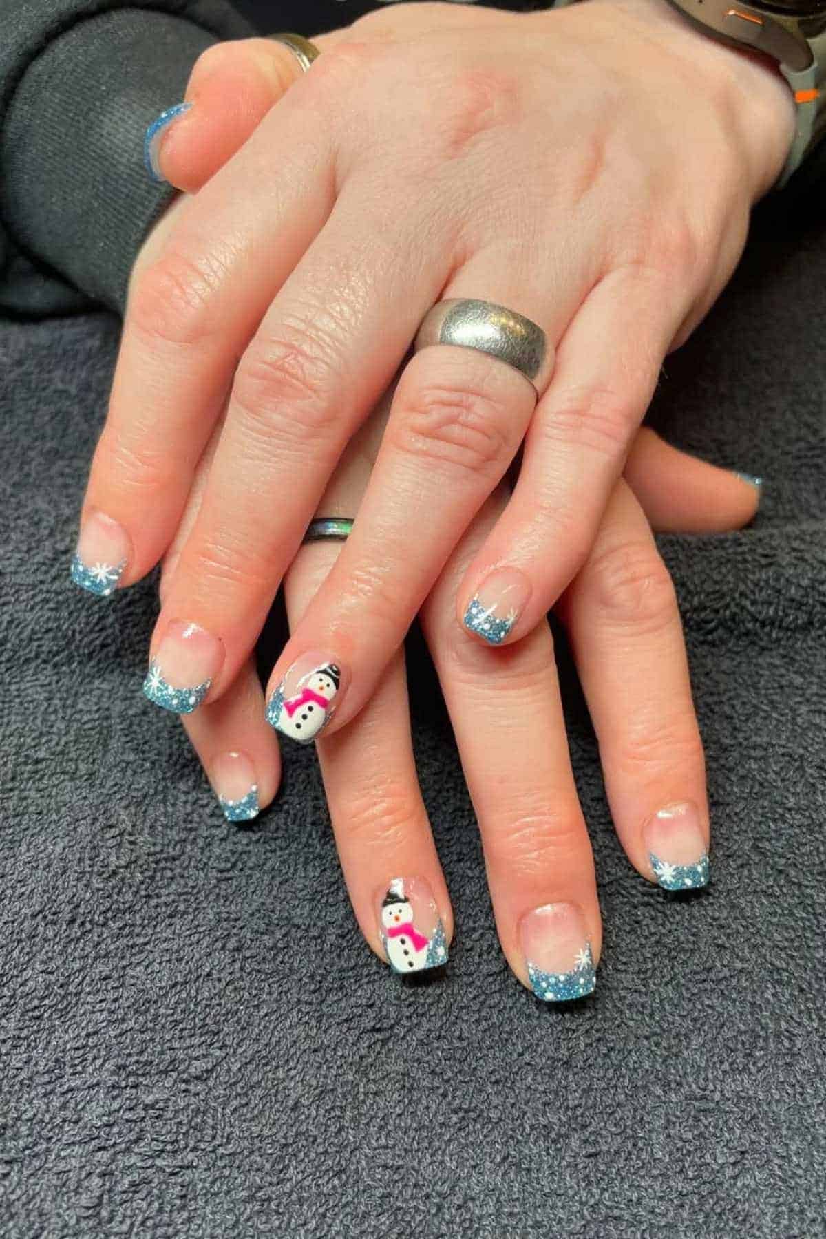Hands with blue glitter French tip nails; two nails feature snowman designs. The person is wearing silver rings on a dark fabric background.