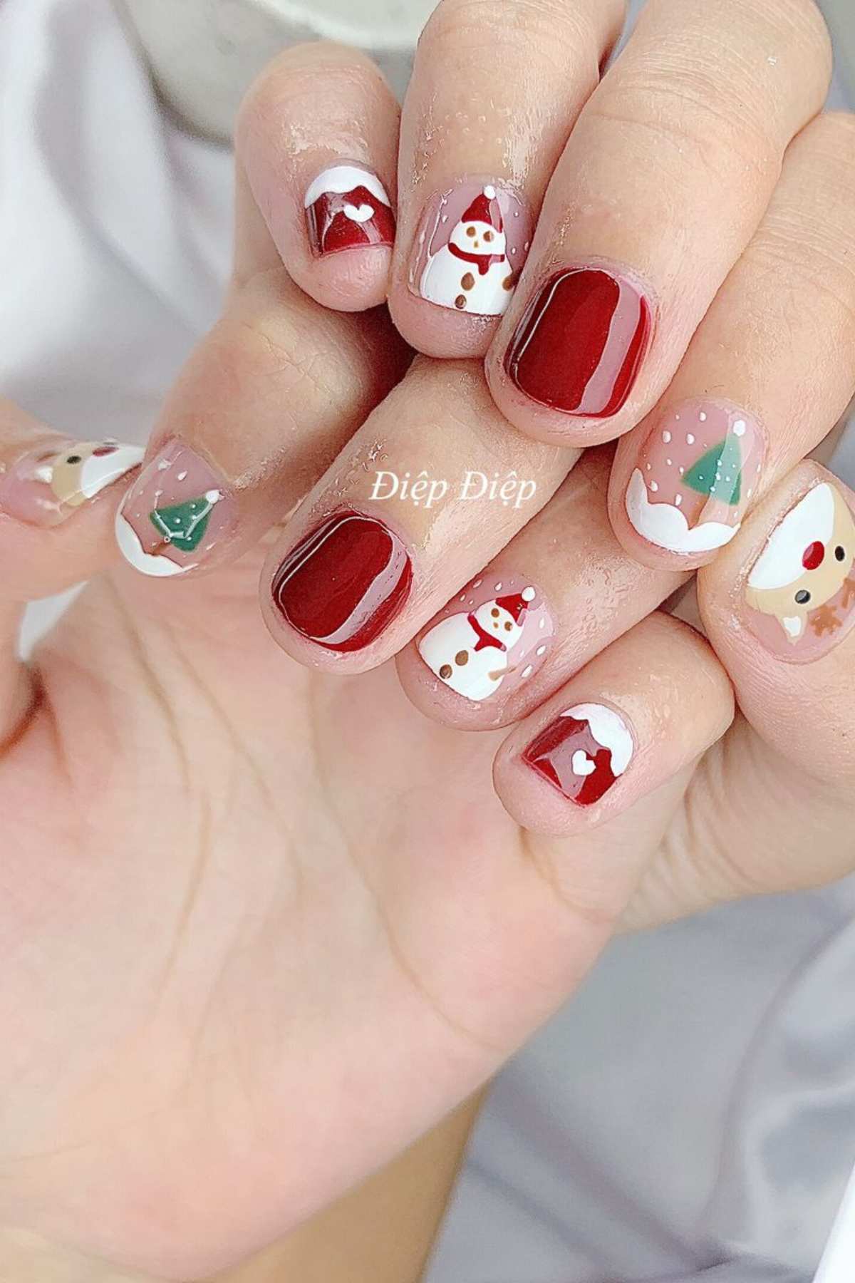 Hands with festive nail art featuring red polish, snowmen, Christmas trees, Santa hats, and reindeer designs on each fingernail.