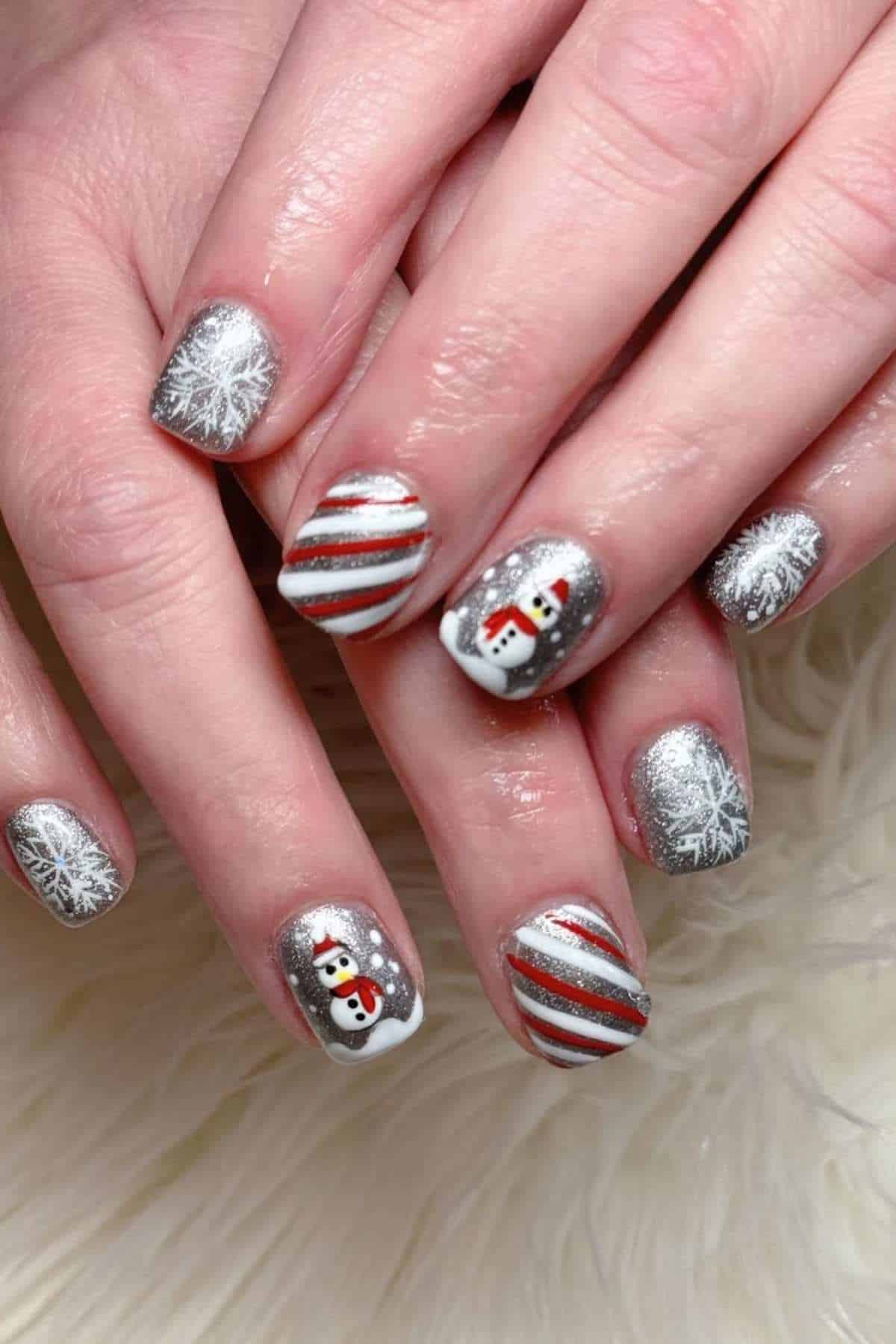 Hands with silver nail polish featuring snowflakes, diagonal red and white stripes, and snowman designs, resting on a white textured surface.
