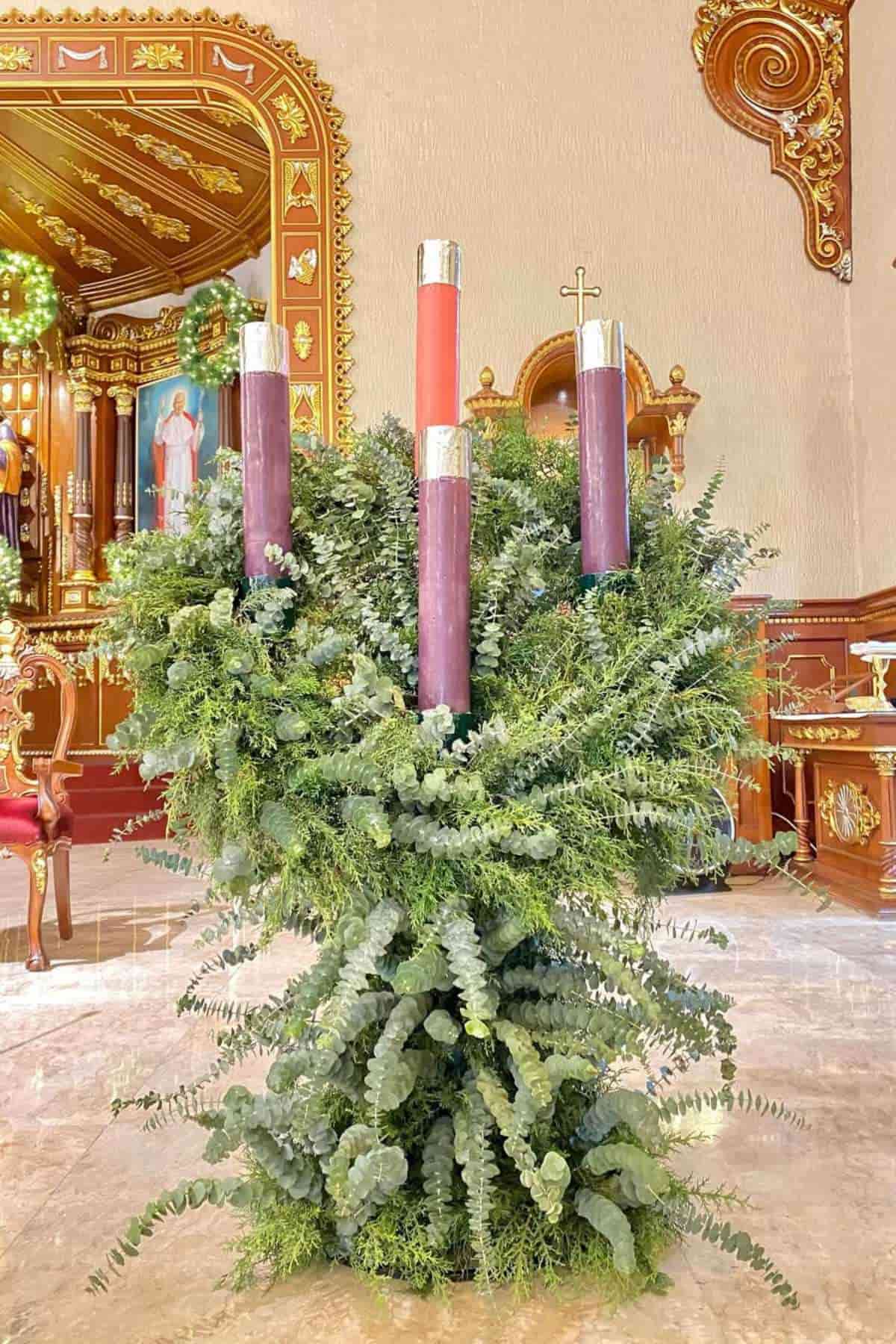A large Advent wreath with three purple candles and one pink candle stands in a church decorated with greenery.