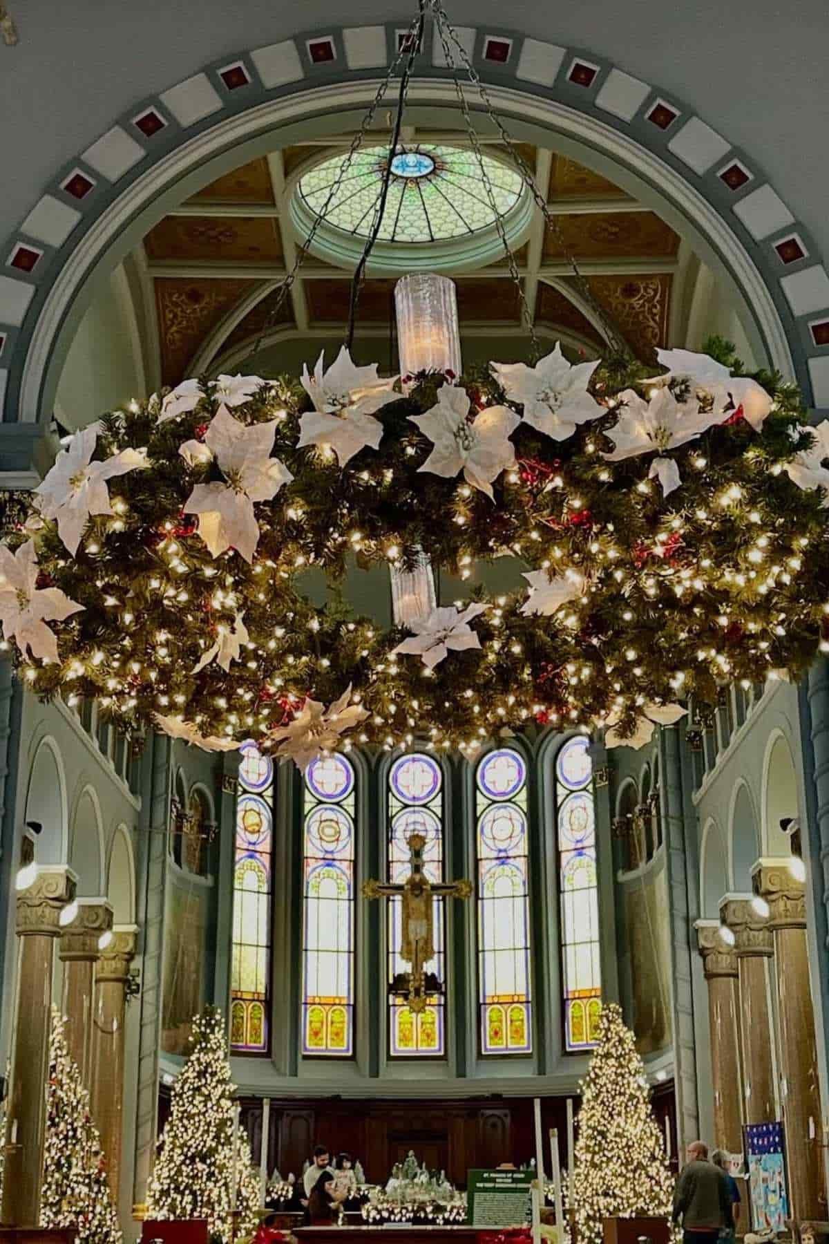 A large, decorated Christmas wreath hangs from the ceiling of a church with stained glass windows and Christmas trees on either side of the altar.