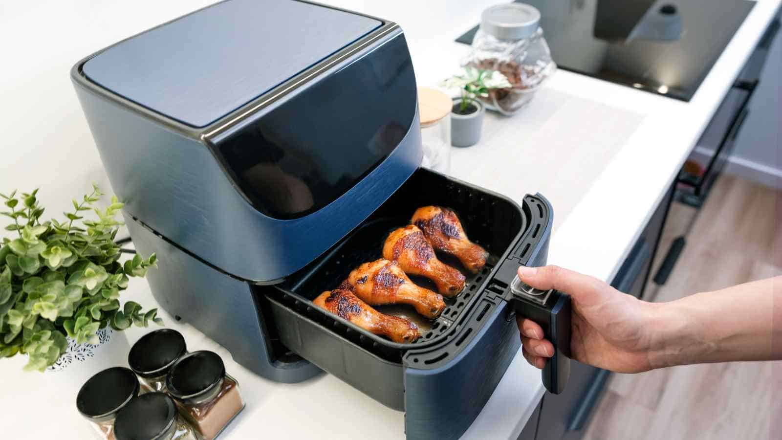 A hand pulls out the basket of an air fryer containing cooked chicken drumsticks in a modern kitchen setting.